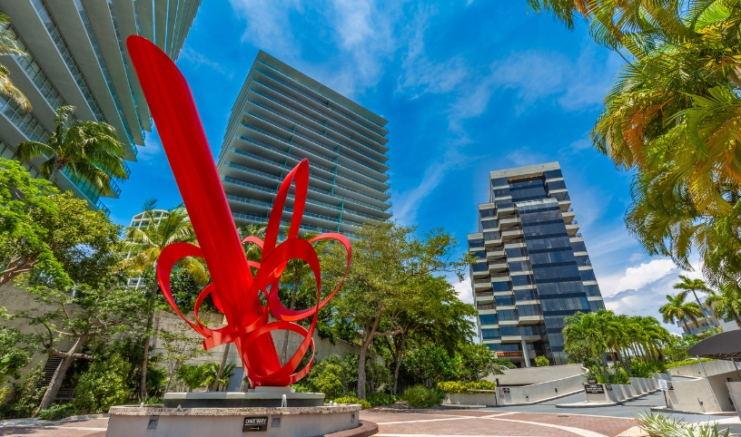 A red abstract sculpture surrounded by palm trees and several buildings.